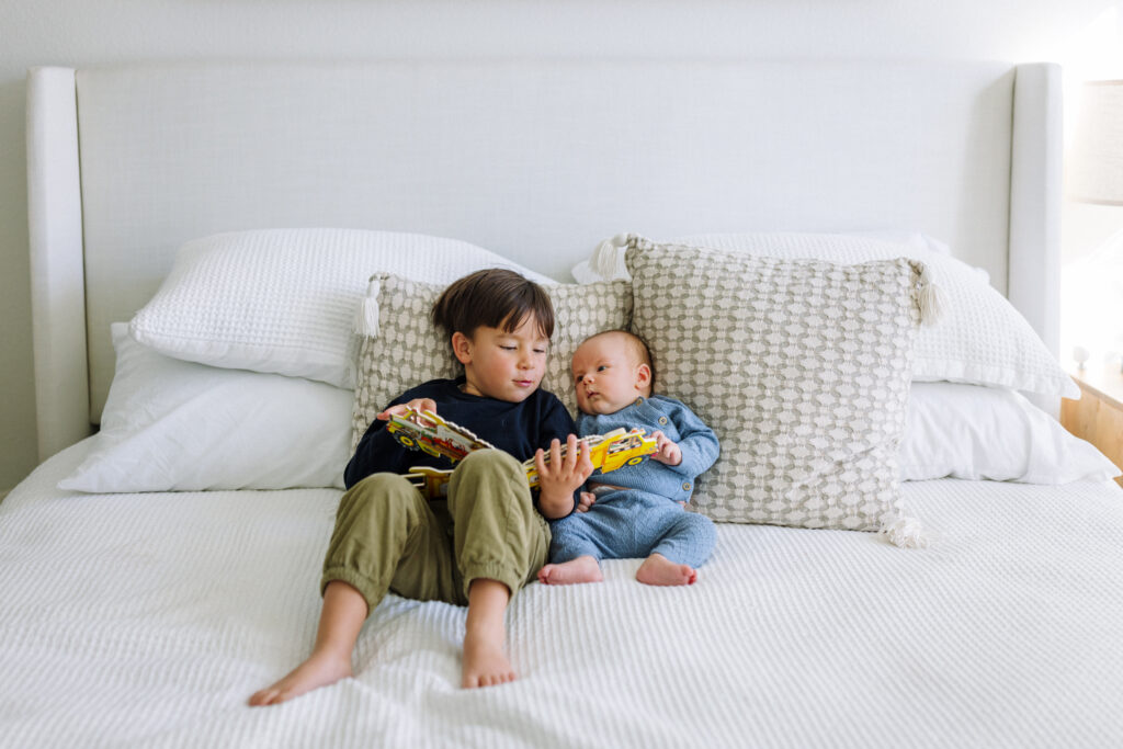 toddler reading book with newborn sibling on bed in-home newborn photography Alameda