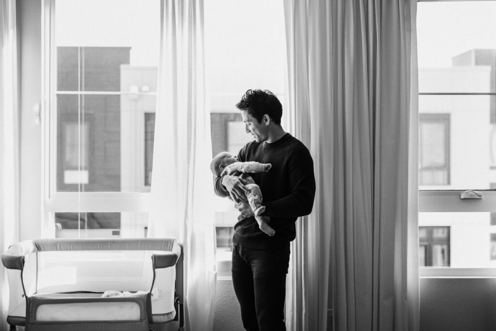 dad holding newborn by window during in-home newborn session Alameda California