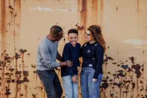 Family of three standing against a rusted orange wall during a family session at the Alameda Naval Base