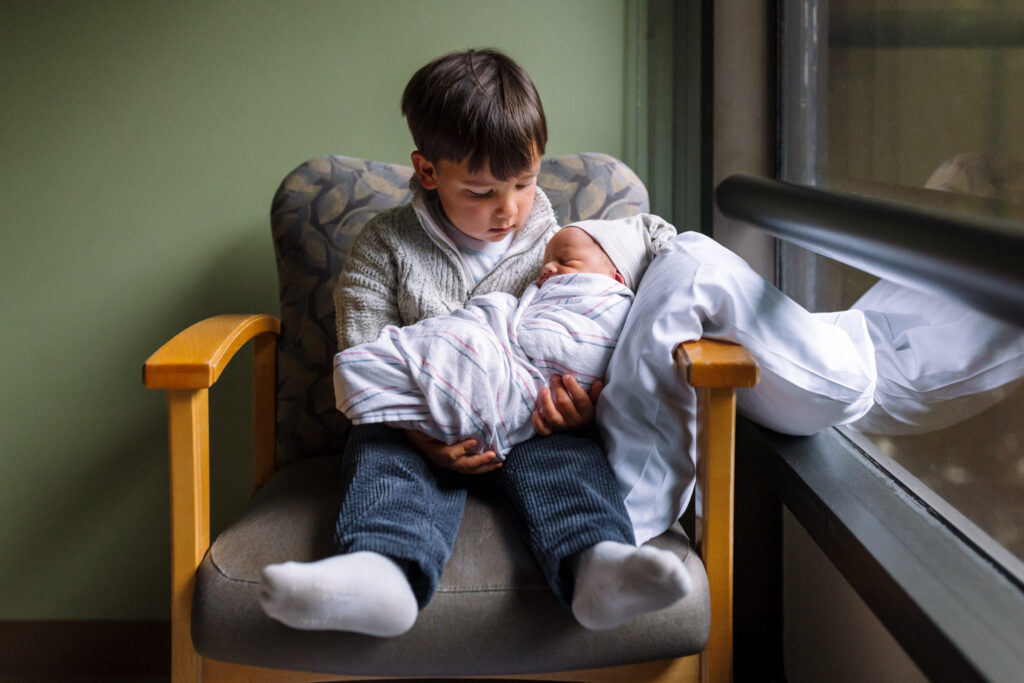 toddler holding newborn baby by hospital window East Bay newborn photographer