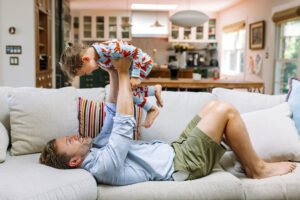 Dad lying on a sofa lifting his daughter in tiger pajamas above him during a playful in-home family session in Oakland
