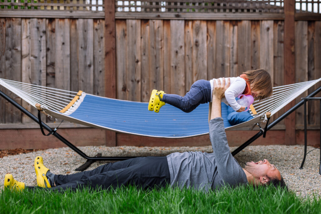 Dad lifting his young daughter above a backyard hammock during a playful outdoor family session in the East Bay