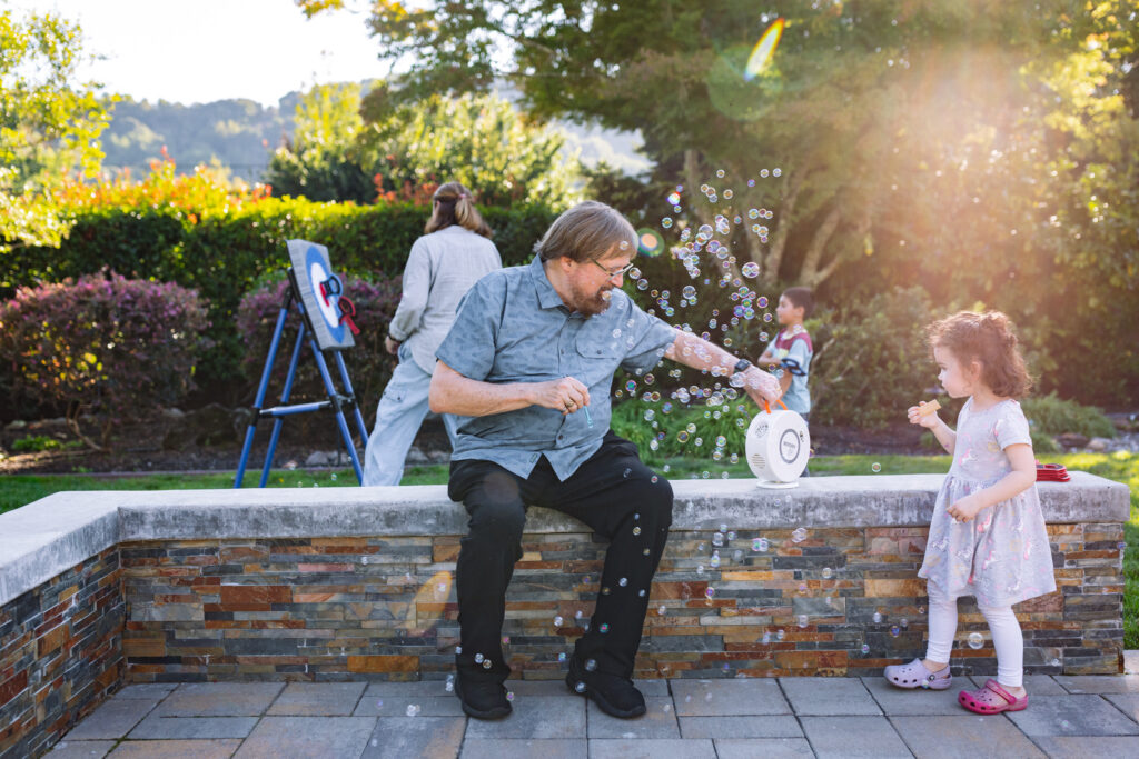 Grandfather blowing bubbles with his toddler granddaughter in the backyard during an outdoor family session in the East Bay