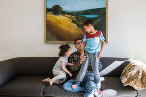 Dad sitting on a couch with his two kids climbing on him during a relaxed in-home family session in Oakland