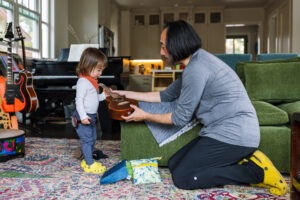 Dad kneeling and showing a ukulele to his toddler in a music-filled living room during an in-home family session in Oakland
