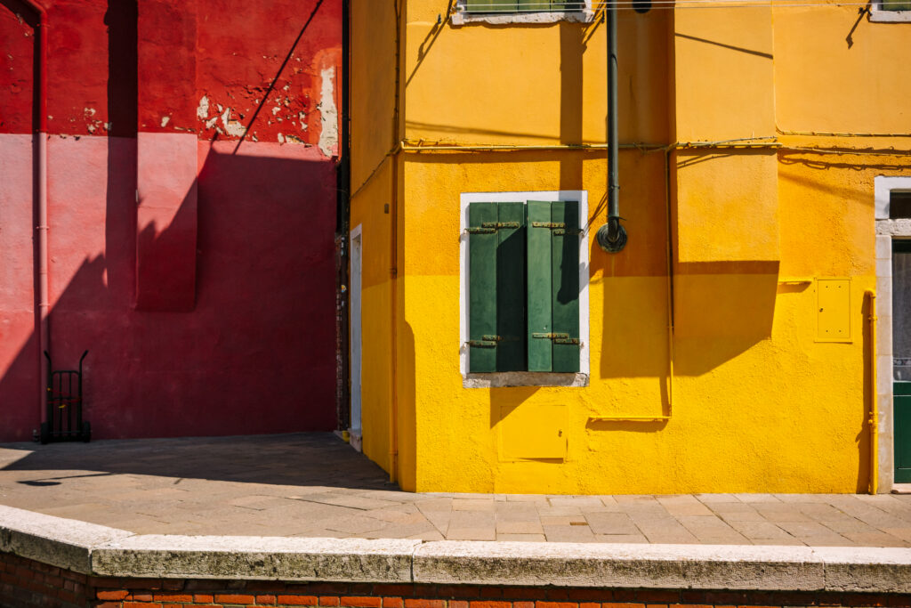 Red and yellow painted facades with green shuttered window and shadows in Burano, Italy