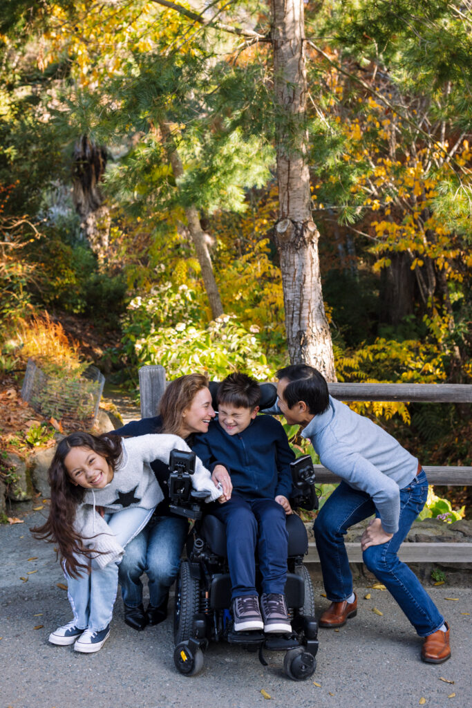 A family surrounds their son in a power wheelchair on the paved path at the UC Botanical Garden in Berkeley, everyone leaning in and laughing together amid fall foliage
