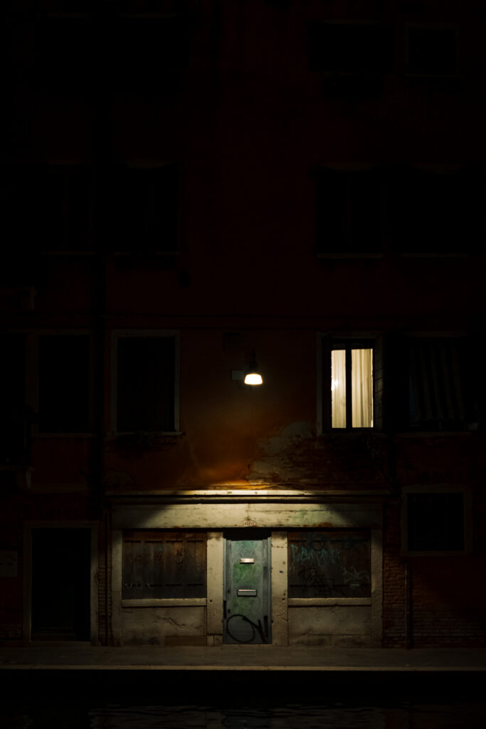 Worn doorway and single lit window on a quiet Venetian street at night