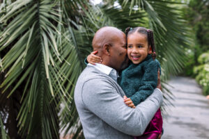 A father holds his young daughter and gives her a kiss on the cheek, both smiling, with large palm fronds behind them at the UC Botanical Garden in Berkeley