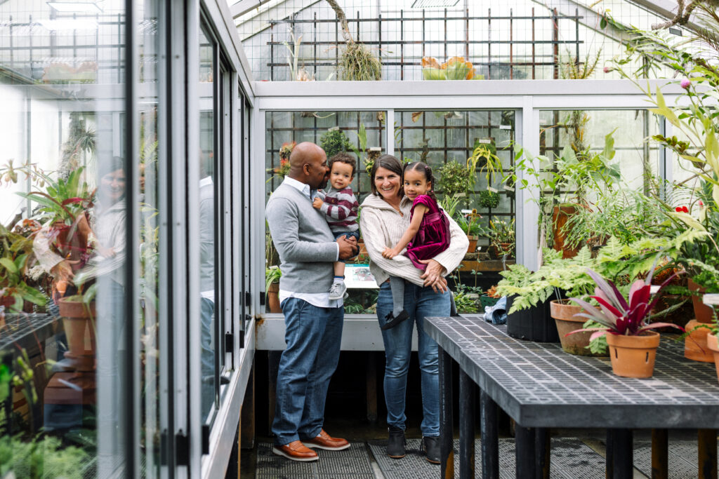 A family of four smiles inside the greenhouse at the UC Botanical Garden in Berkeley, surrounded by tropical plants and natural light