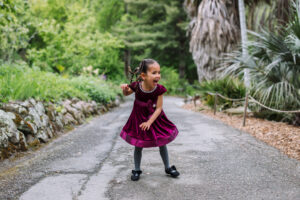 A young girl in a burgundy velvet dress twirls on a paved garden path at the UC Botanical Garden in Berkeley, laughing with her arms out