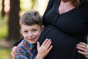 A young boy rests his cheek against his mother's pregnant belly and smiles at the camera in the redwood grove at the UC Botanical Garden