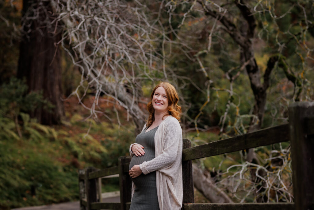 A pregnant woman leans against a wooden garden fence at the UC Botanical Garden in Berkeley, smiling, with dramatic bare branches and fall foliage behind her