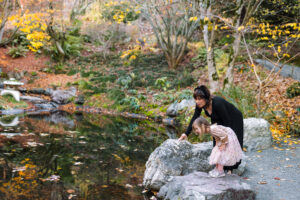 A mother and young daughter crouch beside a pond at the UC Botanical Garden, looking at the water together during a fall family session in Berkeley