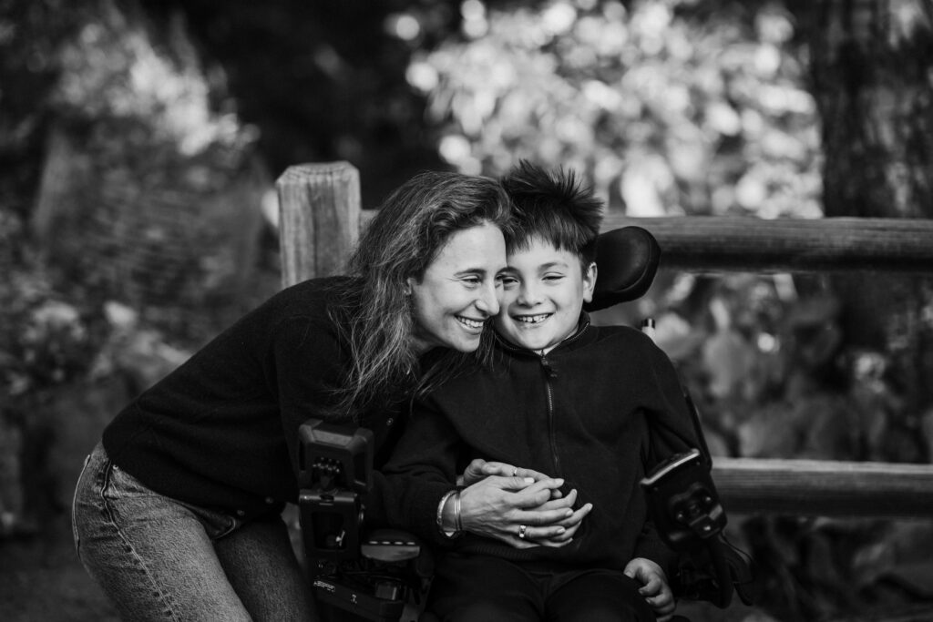 A mother leans in cheek to cheek with her son, who uses a power wheelchair, both laughing during their family session at the UC Botanical Garden in Berkeley