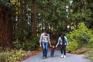 A family of three walks along a paved path near the redwood grove at the UC Botanical Garden in Berkeley, their toddler held between both parents