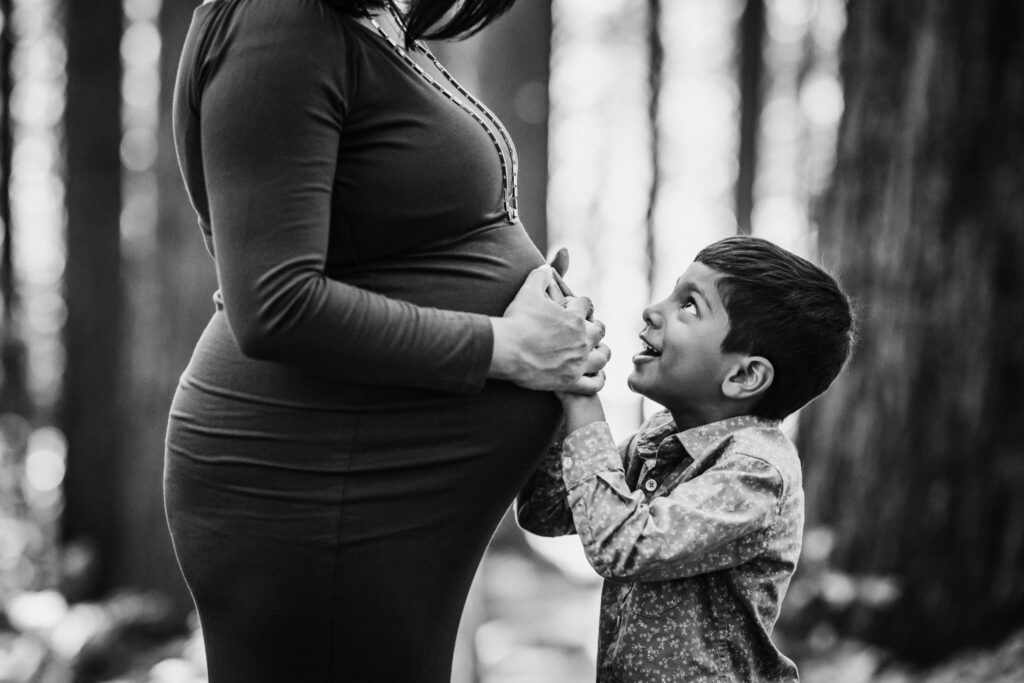 A young boy holds his mother's pregnant belly and looks up at her smiling, surrounded by redwood trees at the UC Botanical Garden