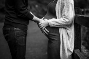 A partner places a hand over a pregnant woman's belly as she holds it gently near a wooden fence at the UC Botanical Garden in Berkeley