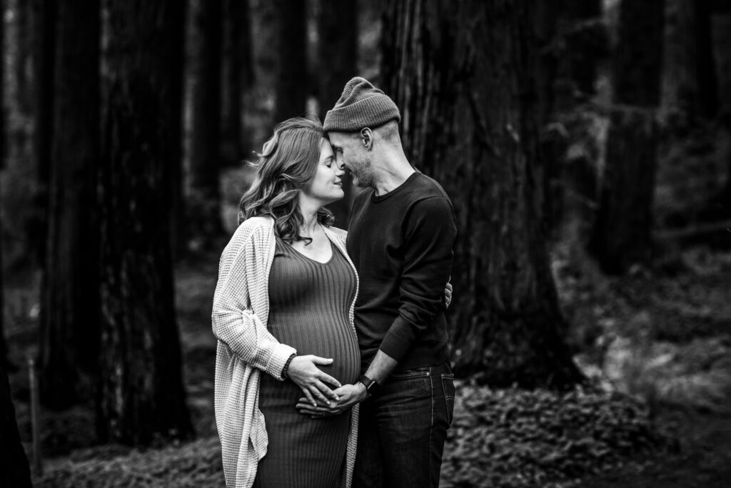 A couple stands close together in the redwood grove at the UC Botanical Garden in Berkeley, foreheads touching, both holding the pregnant belly between them