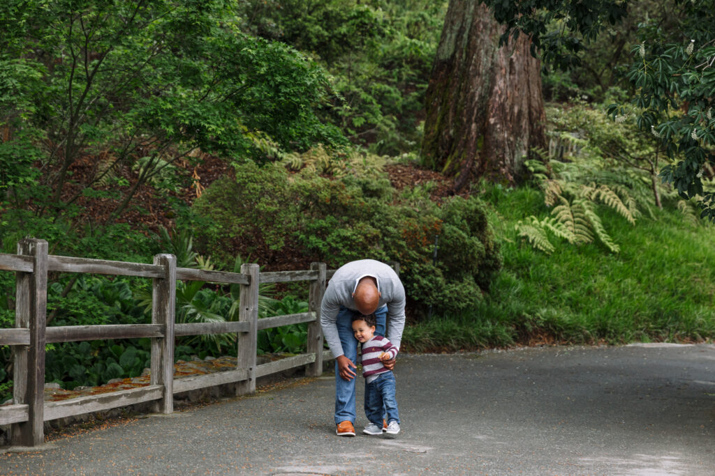 A father bends down to hug his toddler on the paved path at the UC Botanical Garden in Berkeley, with lush greenery and a redwood tree in the background