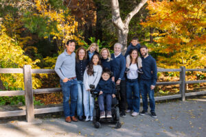 An extended family of ten gathers for a portrait at the UC Botanical Garden in Berkeley, with a child in a wheelchair at the center and vibrant fall foliage behind them