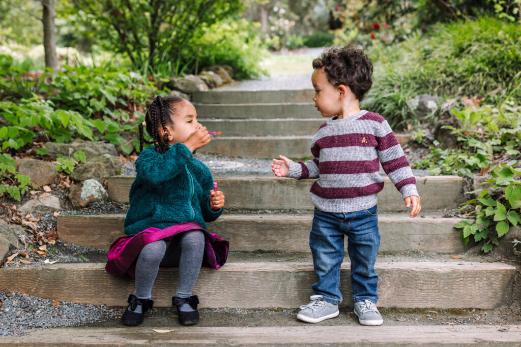 Two young children sit on garden steps at the UC Botanical Garden in Berkeley, one blowing bubbles toward the other