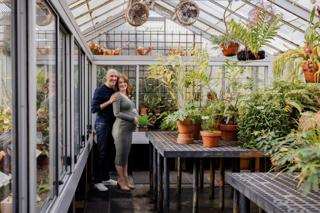 A couple stands inside the greenhouse at the UC Botanical Garden in Berkeley, surrounded by lush tropical plants during a maternity session